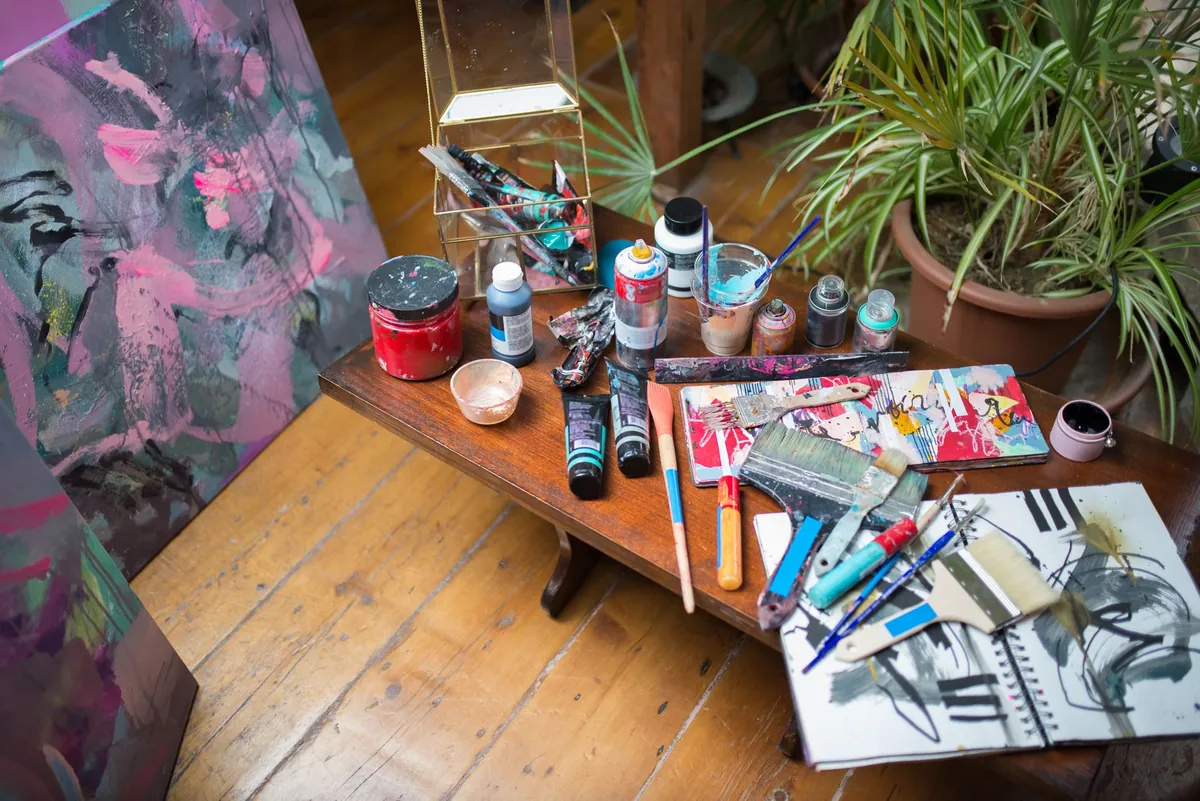 Des tubes de peinture, des pinceaux et des carnets étalés sur une table d'atelier, près de toiles abstraites